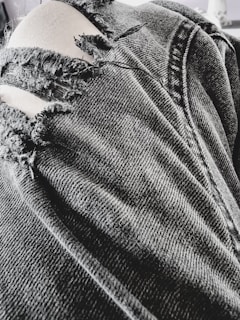 A close-up view of a pair of black, ripped denim jeans with several frayed tears exposing light-colored fabric beneath. The texture of the denim fabric is prominent, showing detailed stitching and wear.