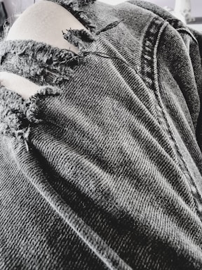 A close-up view of a pair of black, ripped denim jeans with several frayed tears exposing light-colored fabric beneath. The texture of the denim fabric is prominent, showing detailed stitching and wear.