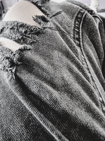 A close-up view of a pair of black, ripped denim jeans with several frayed tears exposing light-colored fabric beneath. The texture of the denim fabric is prominent, showing detailed stitching and wear.