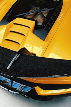 A close-up of the rear part of a sleek, yellow sports car with visible engine components enclosed under a transparent cover. The design is modern and aerodynamic, featuring a bold rear wing with black accents.