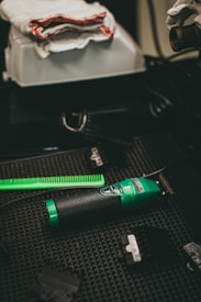 A green and black electric hair clipper lies on a textured dark surface alongside a bright green comb and various clipper attachments. Nearby, a white cloth with red trim rests on a box.