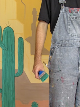 A person wearing paint-splattered denim overalls holds a green paintbrush. The background includes a painted desert scene featuring stylized cacti and a canyon with colors ranging from beige to orange.