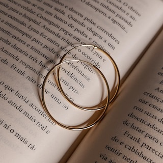 Two thin gold hoop earrings rest on an open book's pages, casting soft shadows. The text on the pages is in a serif font and in Portuguese, detailing a narrative.