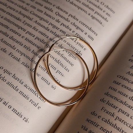 Two thin gold hoop earrings rest on an open book's pages, casting soft shadows. The text on the pages is in a serif font and in Portuguese, detailing a narrative.