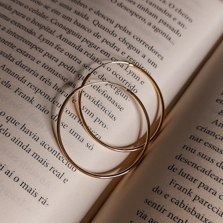Two thin gold hoop earrings rest on an open book's pages, casting soft shadows. The text on the pages is in a serif font and in Portuguese, detailing a narrative.