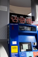A gas station pump displaying prices for three types of gasoline: Regular at $4.29, Extra at $4.69, and Supreme Plus at $4.99. The pump is blue with a digital screen showing the amount of purchase and gallons dispensed. There are various signs and labels on the pump, including a yellow sign indicating alcohol content and text offering disabled access information. The background shows part of the gas station structure and some sunlight and shadows are visible.