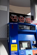 A gas station pump displaying prices for three types of gasoline: Regular at $4.29, Extra at $4.69, and Supreme Plus at $4.99. The pump is blue with a digital screen showing the amount of purchase and gallons dispensed. There are various signs and labels on the pump, including a yellow sign indicating alcohol content and text offering disabled access information. The background shows part of the gas station structure and some sunlight and shadows are visible.