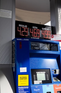 A gas station pump displaying prices for three types of gasoline: Regular at $4.29, Extra at $4.69, and Supreme Plus at $4.99. The pump is blue with a digital screen showing the amount of purchase and gallons dispensed. There are various signs and labels on the pump, including a yellow sign indicating alcohol content and text offering disabled access information. The background shows part of the gas station structure and some sunlight and shadows are visible.