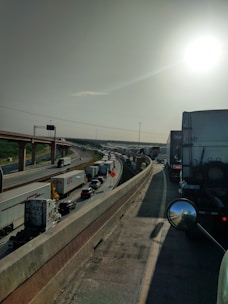 A highway is filled with dense traffic, consisting mostly of large trucks and a few cars. The elevated road curves in the distance and is bordered by concrete barriers. The sun is shining brightly in the sky, casting shadows and creating glare on the vehicles.