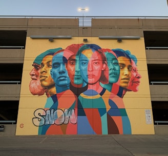 A vibrant mural features overlapping portraits of diverse individuals in a variety of colors. The artwork is painted on the wall of a parking structure and incorporates bright, bold geometric shapes. The word 'SNOW' appears in graffiti style at the bottom left.