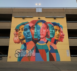 A vibrant mural features overlapping portraits of diverse individuals in a variety of colors. The artwork is painted on the wall of a parking structure and incorporates bright, bold geometric shapes. The word 'SNOW' appears in graffiti style at the bottom left.