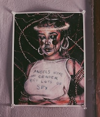 A poster on a wall depicts an expressive, stylized figure with bold lines and contrasting shadows. The figure sports large hoop earrings and appears to have tears resembling paint or ink streaming down their face. They are wearing a sleeveless top with the text 'ANGELS HAVE NO GENDER BUT LOTS OF SEX.' The background features intertwined ropes adding complexity to the composition.