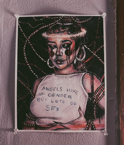 A poster on a wall depicts an expressive, stylized figure with bold lines and contrasting shadows. The figure sports large hoop earrings and appears to have tears resembling paint or ink streaming down their face. They are wearing a sleeveless top with the text 'ANGELS HAVE NO GENDER BUT LOTS OF SEX.' The background features intertwined ropes adding complexity to the composition.