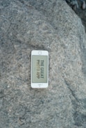 A white smartphone lies on a large, textured rock surface. The phone displays a message on the screen that says 'THE GREAT PHOTO APP'.