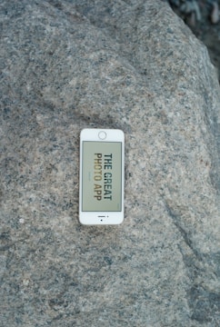 A white smartphone lies on a large, textured rock surface. The phone displays a message on the screen that says 'THE GREAT PHOTO APP'.