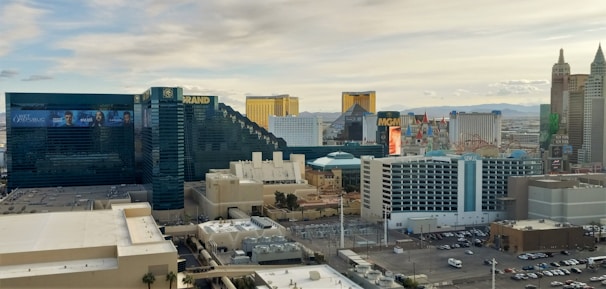 A cityscape view of a bustling urban area featuring numerous large, distinct hotel and casino buildings. Notable structures include the MGM Grand with its recognizable signage, as well as buildings with unique shapes and vibrant architecture that contribute to the city's skyline. A clear sky can be seen in the background, along with distant mountains.