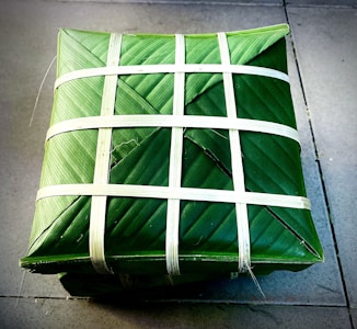 A square package is wrapped meticulously with green leaves and secured with thin white strips woven in a crisscross pattern. The package is placed on a tiled surface, presenting a neat and traditional appearance.