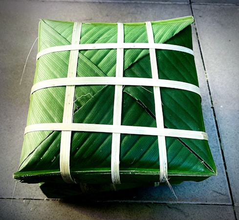 A square package is wrapped meticulously with green leaves and secured with thin white strips woven in a crisscross pattern. The package is placed on a tiled surface, presenting a neat and traditional appearance.