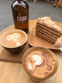 A wooden table holds an arrangement of coffee items and dessert. There is a brown glass bottle labeled 'Cabo Coffee', next to a slice of layer cake on a wooden plate. Two cups of coffee sit in the foreground: one with a latte art design on top.