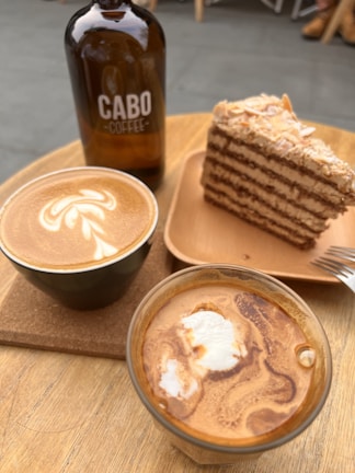 A wooden table holds an arrangement of coffee items and dessert. There is a brown glass bottle labeled 'Cabo Coffee', next to a slice of layer cake on a wooden plate. Two cups of coffee sit in the foreground: one with a latte art design on top.
