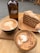 A wooden table holds an arrangement of coffee items and dessert. There is a brown glass bottle labeled 'Cabo Coffee', next to a slice of layer cake on a wooden plate. Two cups of coffee sit in the foreground: one with a latte art design on top.