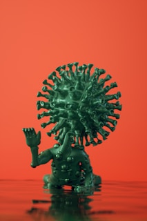 A green humanoid figure with a virus-like head structure sits on a reflective surface against an orange background. The figure has a raised hand and a body covered with spike-like protrusions.