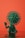 A green humanoid figure with a virus-like head structure sits on a reflective surface against an orange background. The figure has a raised hand and a body covered with spike-like protrusions.