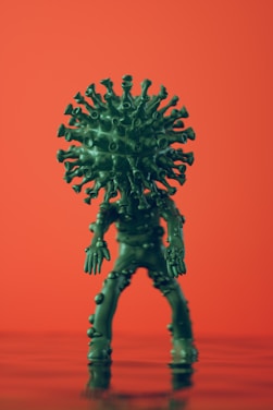 A green, anthropomorphic representation of a virus figure stands against a vibrant orange background. The figure has a spherical head with spike-like protrusions resembling a virus structure and a body covered in smaller nodules. It is posed standing upright with elongated limbs.