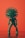 A green, anthropomorphic representation of a virus figure stands against a vibrant orange background. The figure has a spherical head with spike-like protrusions resembling a virus structure and a body covered in smaller nodules. It is posed standing upright with elongated limbs.