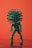 A green, anthropomorphic representation of a virus figure stands against a vibrant orange background. The figure has a spherical head with spike-like protrusions resembling a virus structure and a body covered in smaller nodules. It is posed standing upright with elongated limbs.