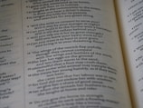 The image features an open book with text printed in a structured column format. The text consists of poetic or prose-like sentences, with numbers indicating sections or verses. The page has a matte finish, and the text is primarily in black ink on an off-white background. The book is slightly angled, showing two pages partially captured in the frame.