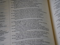 The image features an open book with text printed in a structured column format. The text consists of poetic or prose-like sentences, with numbers indicating sections or verses. The page has a matte finish, and the text is primarily in black ink on an off-white background. The book is slightly angled, showing two pages partially captured in the frame.