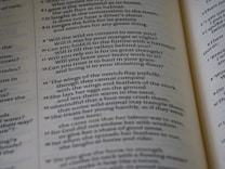 The image features an open book with text printed in a structured column format. The text consists of poetic or prose-like sentences, with numbers indicating sections or verses. The page has a matte finish, and the text is primarily in black ink on an off-white background. The book is slightly angled, showing two pages partially captured in the frame.