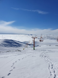 A snowy mountain landscape with ski lifts ascending towards the summit. Tracks and footprints are visible in the snow, suggesting recent activity. The sky is clear with scattered clouds, and a distant view of the mountain range is observed.