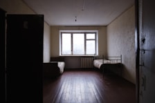 A sparse room with minimal furniture including a single metal bed and a couch, both placed against opposite walls. The room has a large window letting in natural light, and the walls appear bare with a radiator beneath the window. The floor is made of wooden planks, and the lighting is dim.