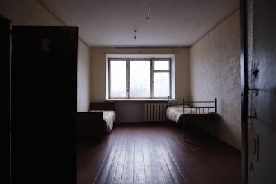 A sparse room with minimal furniture including a single metal bed and a couch, both placed against opposite walls. The room has a large window letting in natural light, and the walls appear bare with a radiator beneath the window. The floor is made of wooden planks, and the lighting is dim.