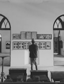 A person stands in an interior space, closely observing a wall with photos and text, possibly in a museum or gallery setting. The architecture includes arched windows and there are several framed images displayed.