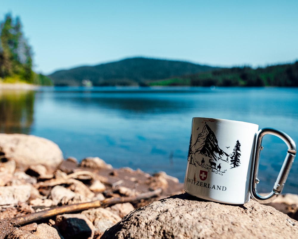 a coffee mug sitting on top of a rock near a lake