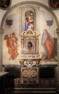 An ornate altar with intricate carvings and a central painting surrounded by decorative elements. The altar is flanked by two painted figures and topped with a stained glass window depicting a vibrant religious scene. The design incorporates classical architectural elements and detailed artwork.