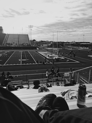 A black and white photograph of a sports field with bleachers in the foreground. A few people sit on the bleachers, and a pair of feet cross over the frame. The field has various markings, goalposts, and a scoreboard. The atmosphere is sparse, with some individuals scattered around the field area. The sky is partly cloudy, creating a moody backdrop.