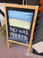 A wooden-framed chalkboard stands on a brick pavement displaying a hand-drawn Ukrainian flag in chalk, with the message 'NO WAR PLEASE' written below it. Several tiles are leaning against a wall in the background, and a fern grows beside the sign.