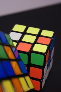 A close-up of two brightly colored Rubik's cubes, placed on a dark surface. One cube appears partially solved with visible green, orange, and yellow faces, while the other is blurred in the foreground.