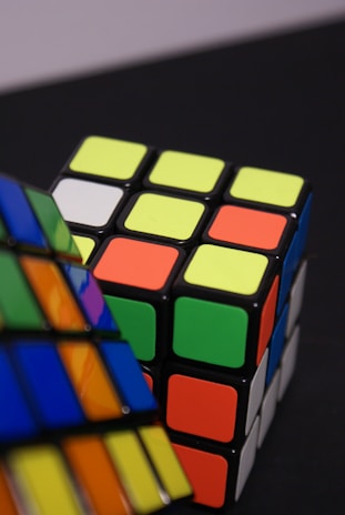 A close-up of two brightly colored Rubik's cubes, placed on a dark surface. One cube appears partially solved with visible green, orange, and yellow faces, while the other is blurred in the foreground.