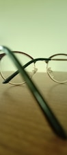 A pair of round, metal-framed eyeglasses is placed on a wooden surface. The background is a blurred pale green, and the focus is on the eyeglasses' frame and temples. Light reflects off the shiny metal parts, creating subtle highlights.