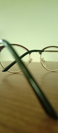 A pair of round, metal-framed eyeglasses is placed on a wooden surface. The background is a blurred pale green, and the focus is on the eyeglasses' frame and temples. Light reflects off the shiny metal parts, creating subtle highlights.
