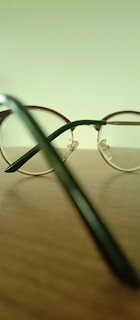 A pair of round, metal-framed eyeglasses is placed on a wooden surface. The background is a blurred pale green, and the focus is on the eyeglasses' frame and temples. Light reflects off the shiny metal parts, creating subtle highlights.