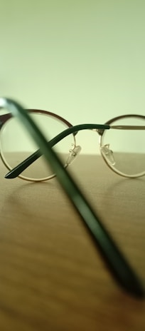 A pair of round, metal-framed eyeglasses is placed on a wooden surface. The background is a blurred pale green, and the focus is on the eyeglasses' frame and temples. Light reflects off the shiny metal parts, creating subtle highlights.