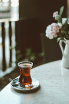 A tulip-shaped glass filled with tea is placed on a saucer atop a marble table. Soft, natural light illuminates the scene, highlighting the translucency of the tea. In the background, a vase with pink flowers and greenery adds a touch of elegance to the setting.