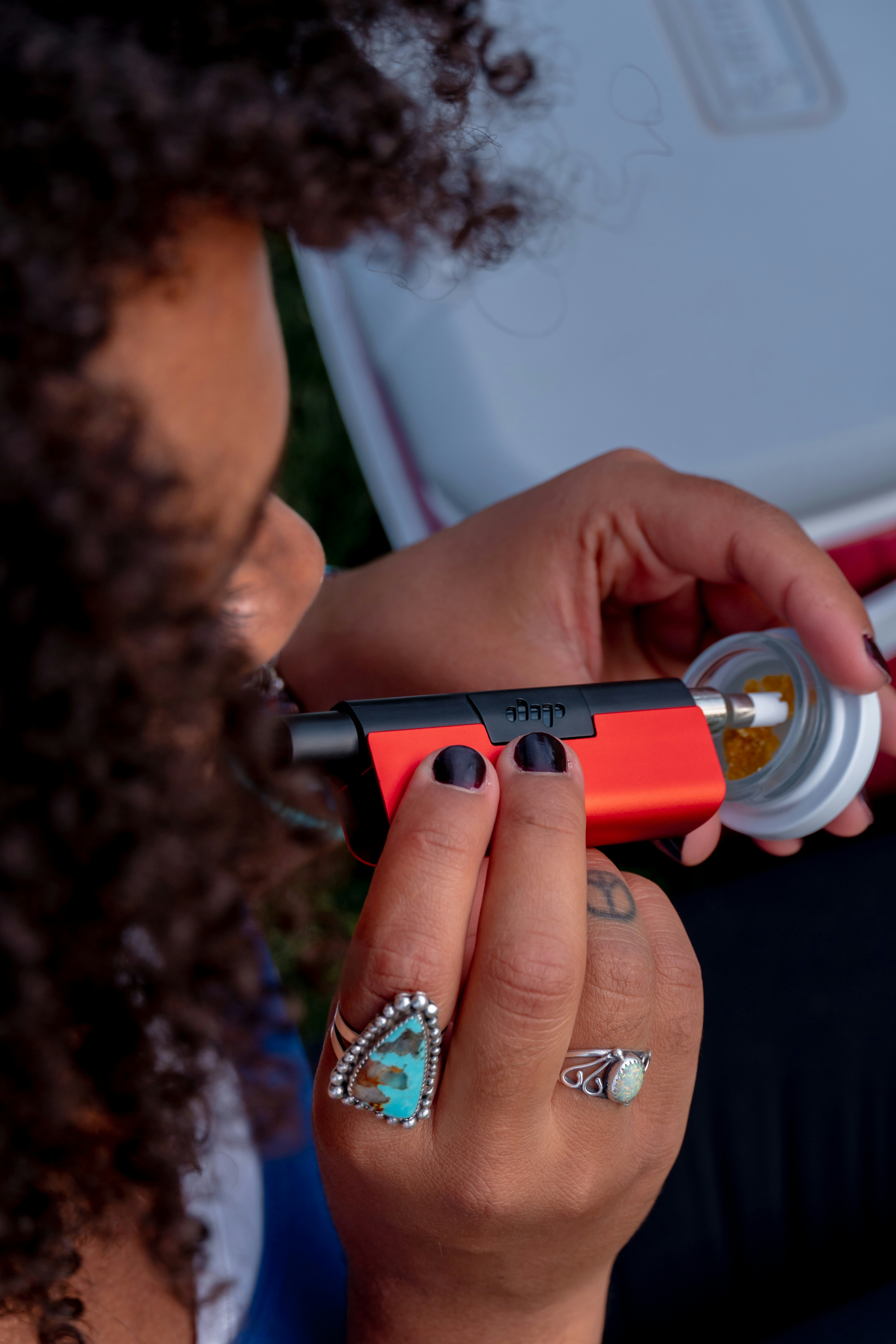 A woman smokes using a red Dip Devices EVRI 3-in-1 dab and vape concentrate device. She has curly hair, black nail polish and is wearing fashionable rings. See more at https://dipdevices.com/