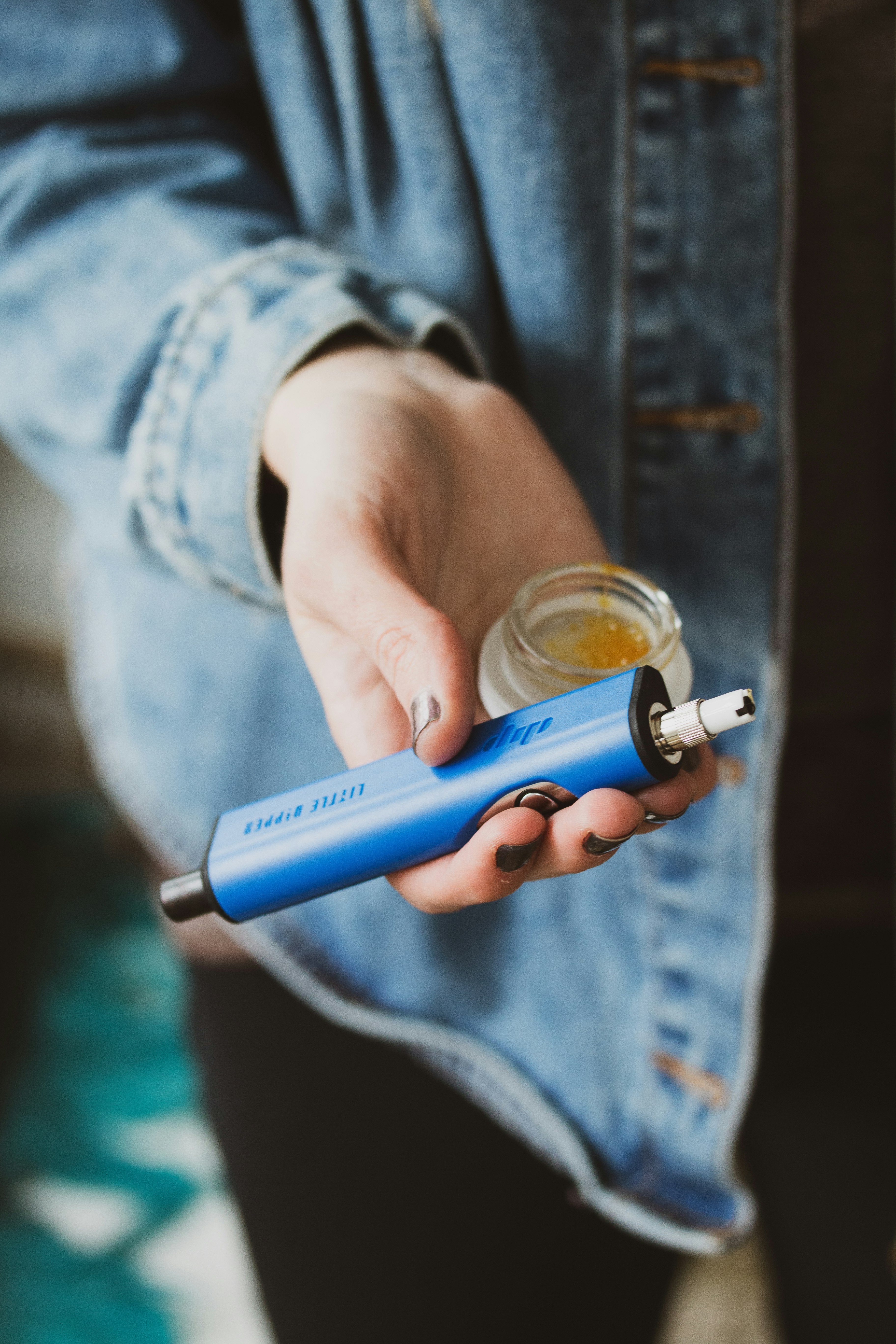 A person holds a blue Dip Devices Little Dipper concentrate vaporizer dab straw as if offering it. They wear metallic nail polish and also hold a container of cannabis extract.

See more at https://dipdevices.com/ 